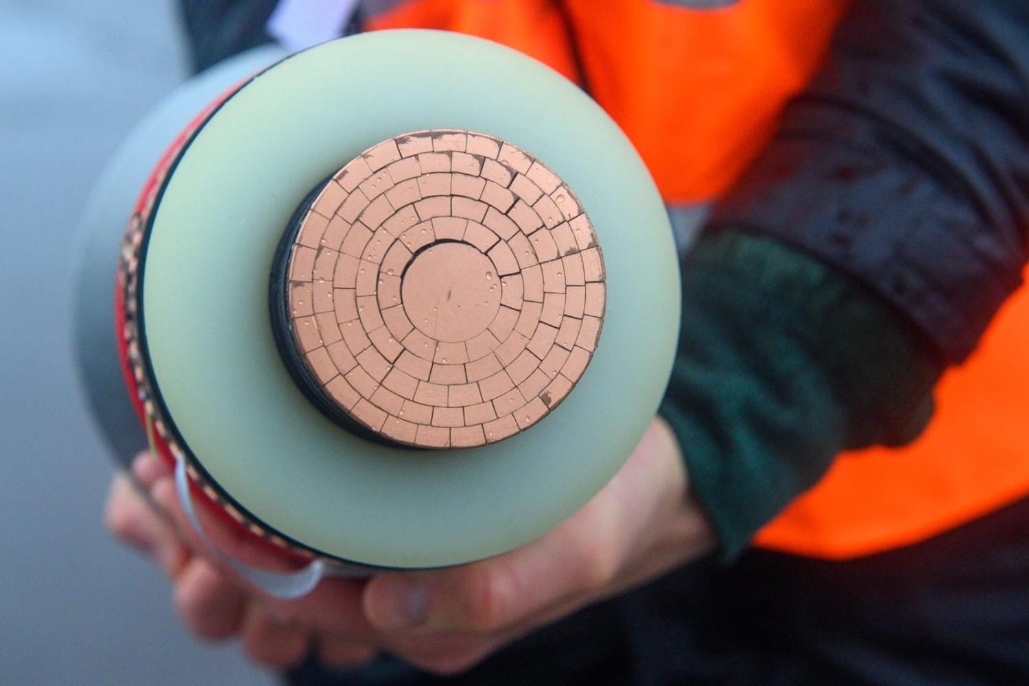 A person wearing an orange safety vest holds a cross-section of a high-voltage power cable, showing multiple concentric copper segments.