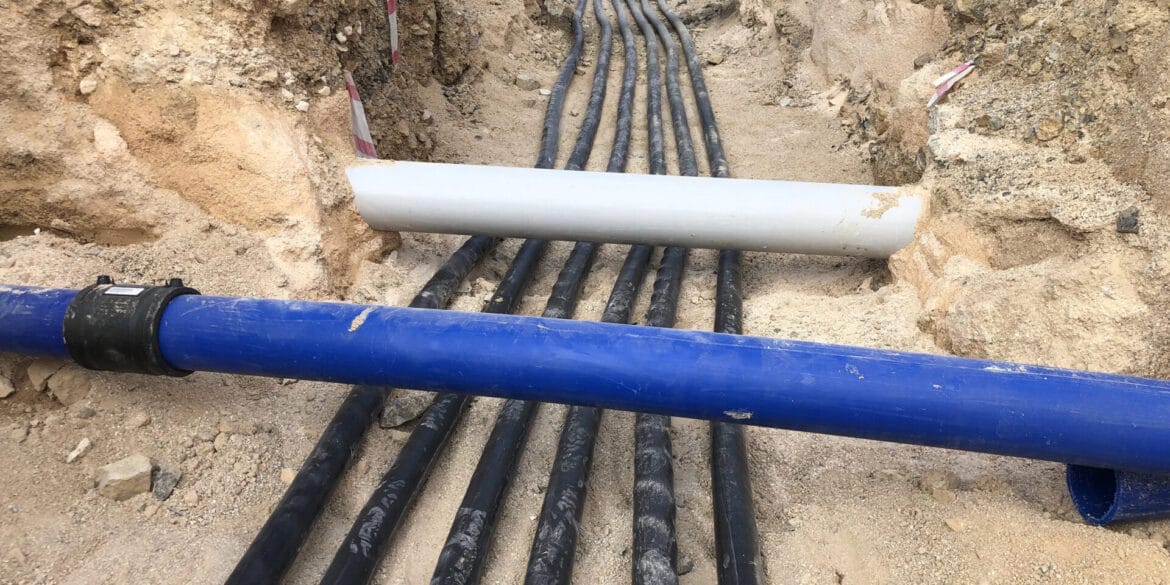 Multiple black cables and two large pipes, one blue and one white, run through a trench dug in sandy soil at a construction site.