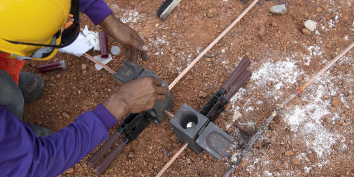 Worker in a yellow helmet prepares electrical grounding connections on copper wires using welding molds and tools on a dirt surface.