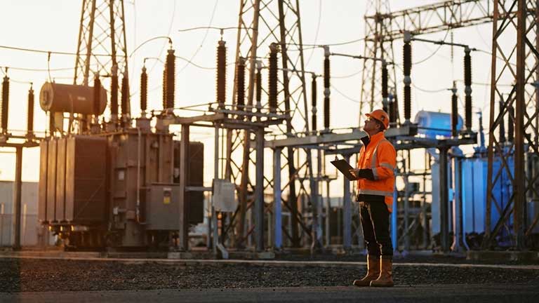 A worker in a high-visibility jacket and helmet stands with a tablet at an outdoor electrical substation surrounded by power lines and equipment.