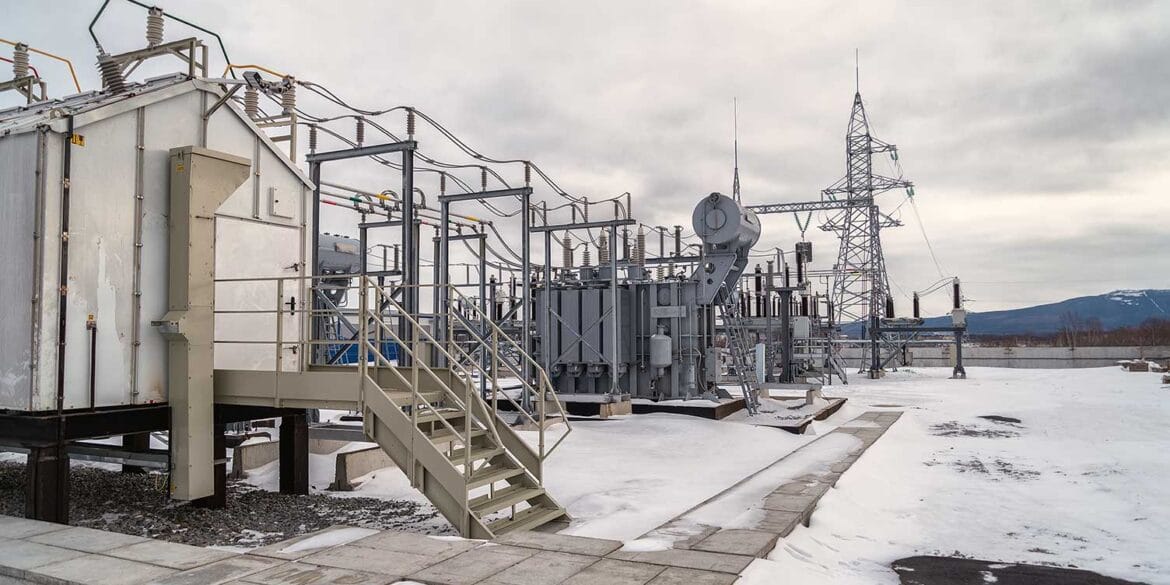 Snow-covered electrical substation with transformers, metal structures, and power lines under a cloudy sky; mountains visible in the background.
